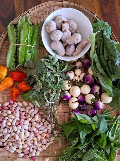 A basket of fresh ingredients ready for cooking. It includes winged beans, taro, Raja Mircha chillies, kidney beans, bitter eggplant, and fresh mustard greens.