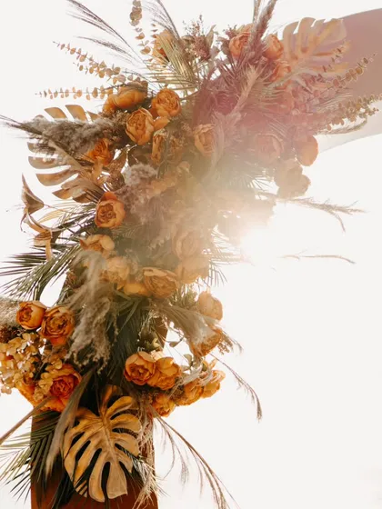 A close-up of a rustic floral arrangement featuring pampas grass and dried leaves. The sun flare adds a dreamy, golden-hour touch to this boho-chic decor detail.