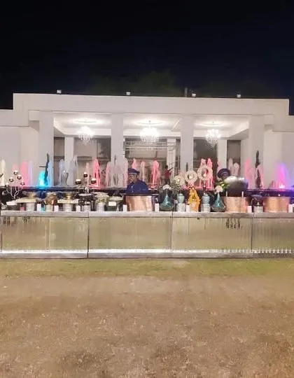 A spectacular night-time buffet setup in front of a large fountain with colorful lights. The long, illuminated counter adds a dramatic and festive feel to this large outdoor wedding reception.