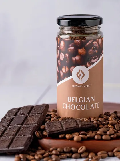 A clean product shot focusing on a jar of Belgian Chocolate instant coffee. The composition uses real chocolate and coffee beans as props to emphasize the product's core ingredients.