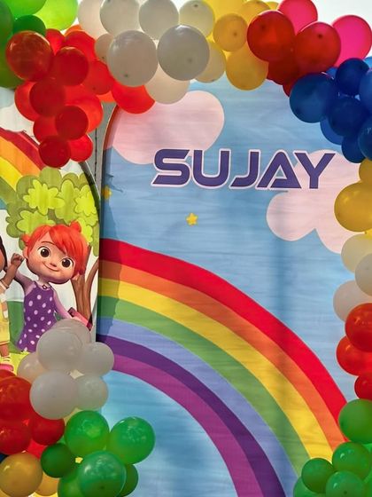 A bright and cheerful Cocomelon-themed first birthday party. The setup features a character backdrop and a rainbow-colored balloon arch.