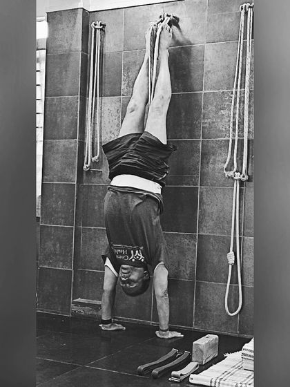 A black and white photo of a handstand (Adho Mukha Vrksasana) using the wall ropes for support. The ropes help to achieve vertical alignment and build shoulder stability.