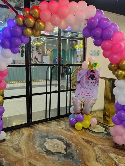 A colorful balloon arch at the entrance of a venue for a first birthday party, with a personalized welcome sign.