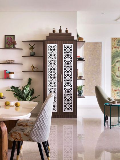 This dining area combines modern and traditional elements. The sleek marble-top table with a detailed wooden base is paired with upholstered chairs. In the background, a puja unit with jali-patterned doors adds a touch of Indian heritage.