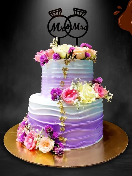 This two-tier engagement cake features a beautiful purple and white ombre buttercream finish. I decorated it with a cascade of fresh flowers and a touch of edible gold leaf for a modern and romantic look.