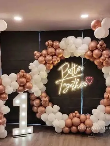 A 'Better Together' first-anniversary setup with a circular balloon arch in rose gold and white, and a '1' marquee light.