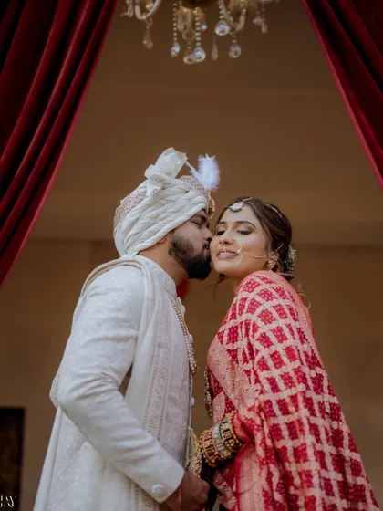 A close-up capturing a sweet kiss on the cheek. This intimate moment is beautifully framed by the rich red curtains, highlighting the couple's joy and affection.
