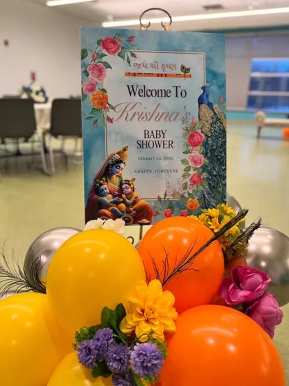 The welcome easel for a 'Krishna's Baby Shower' event. The sign is surrounded by a vibrant arrangement of orange and yellow balloons and flowers, setting a festive tone right from the entrance.