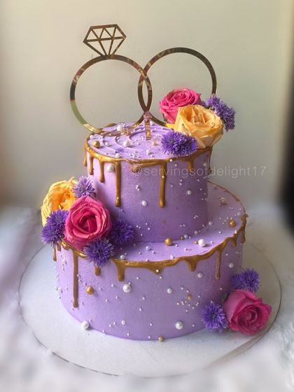 A vibrant two-tiered engagement cake in a beautiful purple hue. It's decorated with a gold drip, fresh flowers, and a classic ring topper to celebrate the special moment.