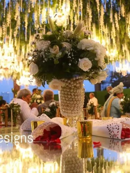 A close-up of a guest table under a canopy of hanging flowers and lights. The reflection on the mirrored tabletop creates a beautiful, layered effect, doubling the magic of the decor above.