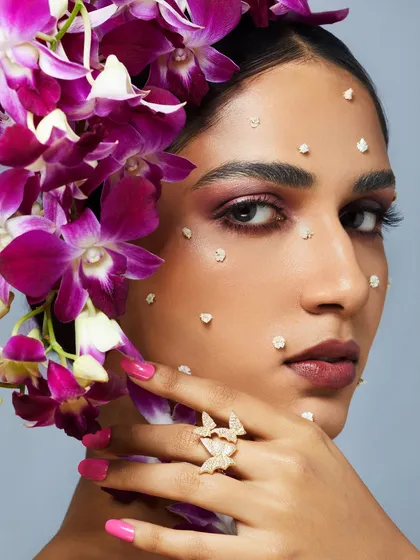 A soft, romantic makeup look with purple tones, accented by real orchids and delicate butterfly-like details on the skin.