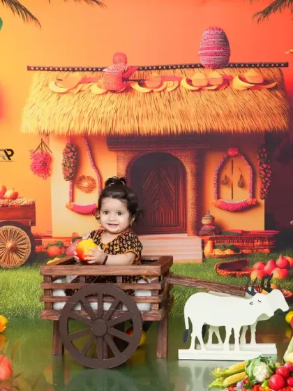 A baby sits in a wooden cart in a rustic village or farm-themed setup. The warm colors and traditional props like the hut and cow figures create a charming and nostalgic scene.