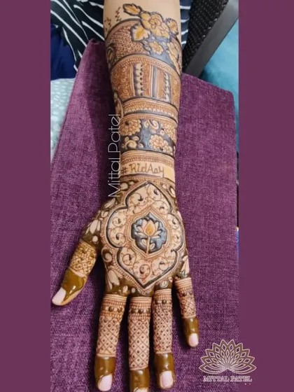 A detailed shot of a bridal back of hand design. The central lotus motif is surrounded by intricate patterns, and the arm features a floral band and personalized names.