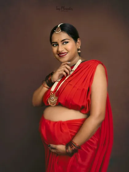 The vibrant red saree and traditional jewelry make for a striking and joyful portrait, celebrating motherhood with cultural richness.