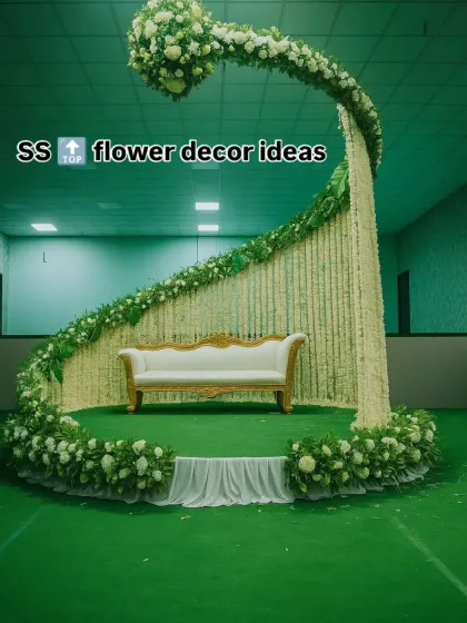 A unique and modern stage design featuring a large, curved structure made of white flowers and greenery against a green backdrop. The minimalist white sofa adds to the contemporary and fresh feel of this indoor setup.
