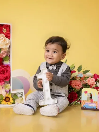 A beautiful first birthday setup with a floral number '1' and bouquets of flowers. The baby boy, dressed in a smart vest, holds a light-up number one, adding a touch of magic to the scene.