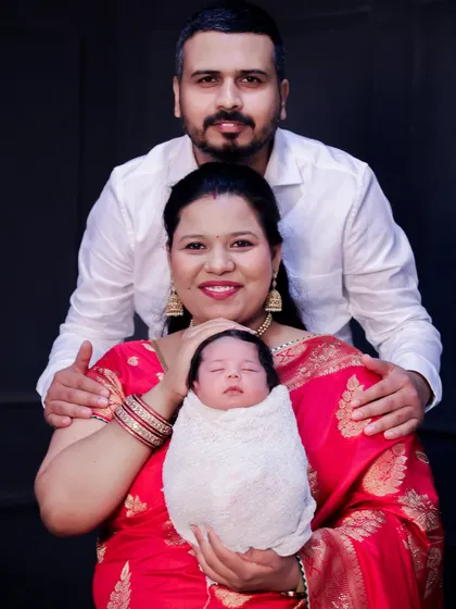 A lovely portrait featuring a mother in a vibrant red saree holding her newborn, with the father standing protectively behind them. It’s a wonderful mix of traditional attire and modern family photography.