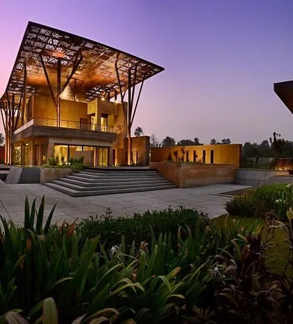 The Tree House at dusk, set within its landscaped garden. The design includes multiple outdoor zones, including a large stone plaza and a sculptural pavilion, extending the home's living spaces into the open air.