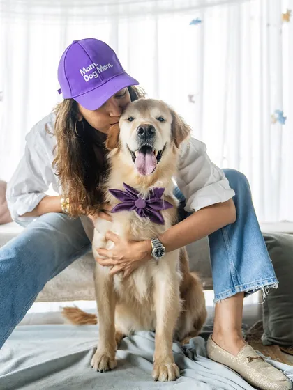 A proud dog mom wearing a "Dog Mom" cap gives her Golden Retriever a kiss. The matching purple bow on the dog adds a touch of coordinated cuteness.