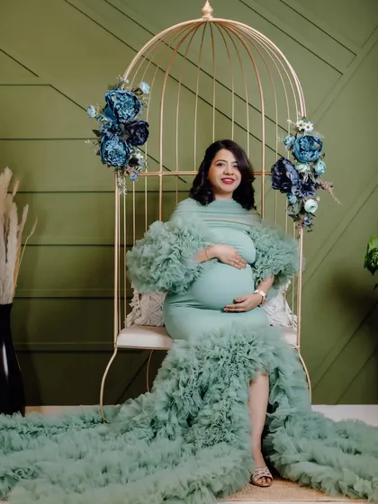 This mom-to-be looks absolutely serene in a mint green mermaid-style maternity gown. The studio setup with the golden birdcage chair and floral decor creates a beautiful, elegant portrait.