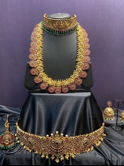 A classic temple jewellery set with a long kasulaperu (coin necklace) and a matching choker. The red and green kemp stones add a vibrant touch to the antique gold finish.