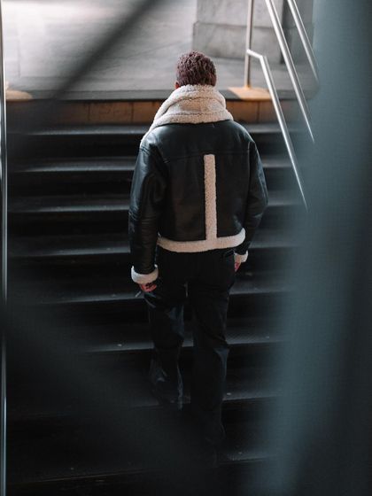 A moody shot taken in a New York subway station. The framing through the foreground railing and the dark tones emphasize the texture of the shearling jacket and create a sense of urban solitude.