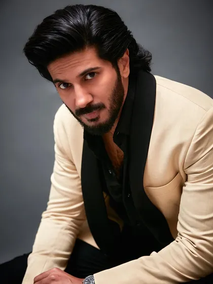 A close-up portrait of Dulquer Salmaan from the GQ shoot. The focus is on his expressive look, framed by the sharp contrast of the cream and black formalwear.