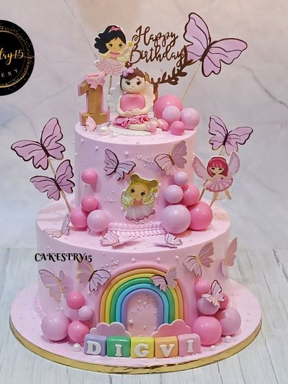 A mesmerizing two-tier cake with a fairy enchantment theme for a first birthday. It's decorated with fondant fairies, a rainbow, and fluttering butterflies in pastel hues.