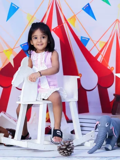A sweet two-year-old girl sits on a rocking horse in front of a vibrant carnival backdrop. A classic and playful birthday portrait.