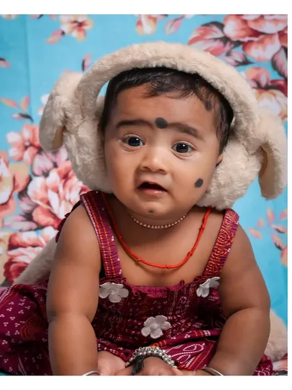 A close-up portrait of a baby girl wearing adorable, fluffy earmuffs. Her wide, curious eyes and the vibrant floral background make this a truly charming and memorable shot.