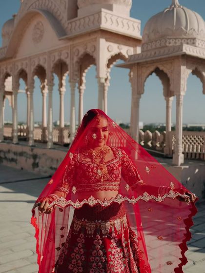 The bride playfully peeks through her vibrant red dupatta, her smile radiant against the backdrop of a royal Rajasthani palace.