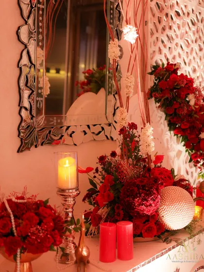 A close-up of a decor console, where an ornate mirror reflects the warm, candlelit ambiance, surrounded by lush arrangements of red roses and decorative spheres.