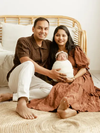 A happy family of three posing for a classic portrait. The parents are smiling at the camera while holding their precious newborn, dressed in coordinating earthy tones.