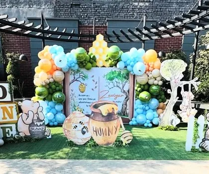 An enchanting outdoor Winnie the Pooh first birthday party, designed as an open storybook. This wide shot shows the full setup on a green lawn, with Pooh and friends character cutouts, a "Hunny" pot prop, and colorful balloon arrangements, creating a whimsical scene straight out of the Hundred Acre Wood.