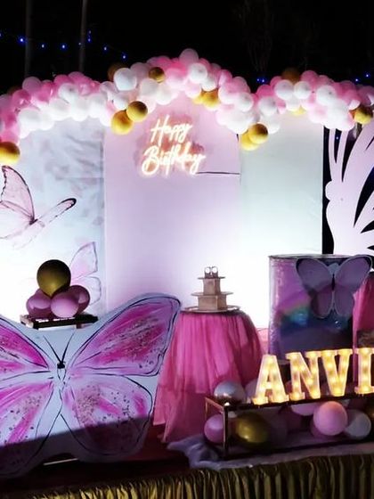 An evening shot of a butterfly-themed birthday for Anvika. The lighting highlights the large butterfly wing props and the personalized marquee name sign, creating a dramatic and enchanting atmosphere for the party.
