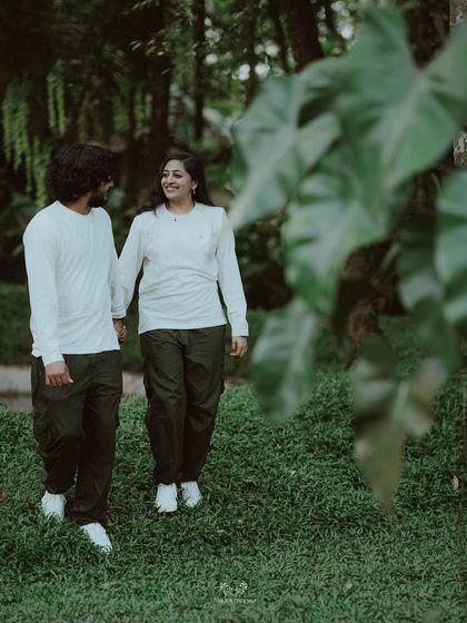 The couple walking hand-in-hand through a wooded path, framed by lush green leaves.