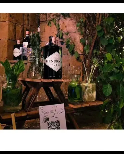 A rustic gin bar setup nestled amongst natural greenery. This 'garden bar' concept uses wooden stands and glass jars filled with fresh herbs and botanicals for a truly organic feel.