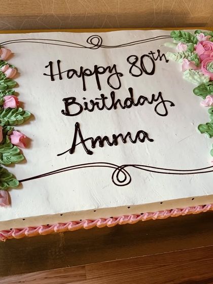 A beautiful sheet cake for an 80th birthday celebration for "Amma." The cake is bordered with hand-piped pink buttercream roses and green leaves, a classic design filled with love.