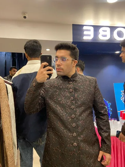 A customer's mirror selfie while trying on a dark, subtly printed Indo-western sherwani in our store. Tradition meets modern style.