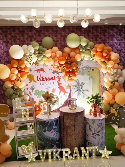 A fun and friendly dinosaur theme perfect for a young explorer's first birthday. The setup uses an earthy color palette of orange and green balloons, a custom-printed dinosaur backdrop, and cute dinosaur cutouts.