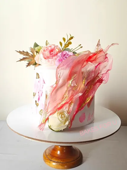 A modern cake featuring a dramatic, flowing rice paper sail in shades of pink and gold. This artistic element adds height and movement to the design, complemented by fresh flowers.