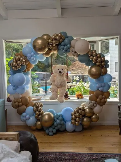 A beautiful teddy bear themed balloon ring set against a window. The arrangement in blue, brown, and gold is perfect for a baby boy's celebration.