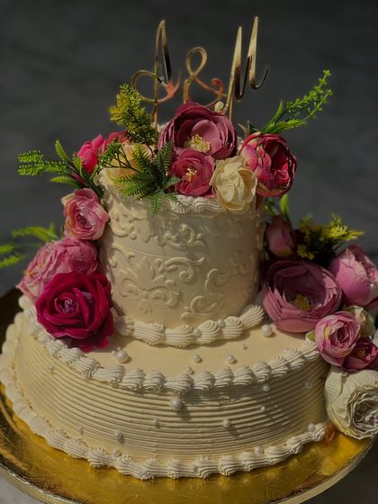 This two-tiered cake features intricate stencil work and is adorned with beautiful pink and white flowers. The custom initial topper adds a personal touch for the couple's big day.