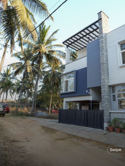 The modern exterior of the Shashidhar Residence, set against a backdrop of coconut trees. The design uses a simple palette of white, grey, and stone to create a clean and contemporary look.