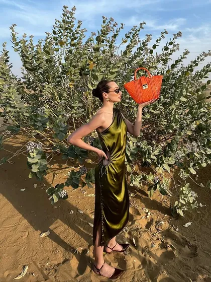 A behind-the-scenes shot from the Da Milano desert campaign. This image captures the model in a one-shoulder satin dress, holding a vibrant orange mini-bag against the backdrop of desert flora, showcasing the on-location styling process.