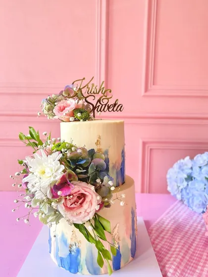 This two-tiered engagement cake features a dreamy blue and pink watercolor finish, gold leaf accents, and a cascade of fresh flowers.