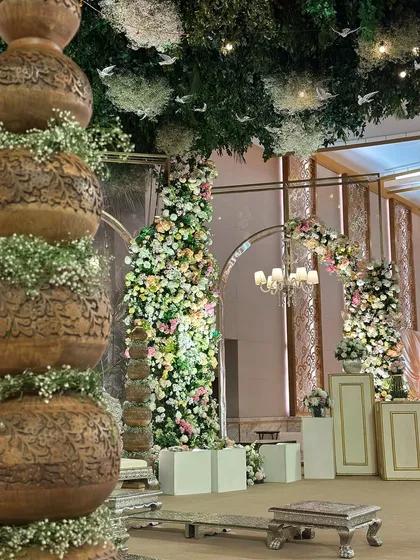 A detail shot of the vintage garden decor, featuring traditional carved pots and towering floral installations. The ceiling is a canopy of green leaves and hanging white birds.