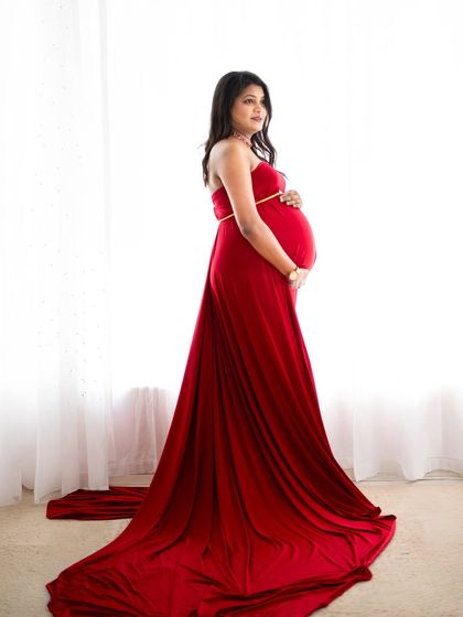 A classic and elegant silhouette shot. The mom-to-be stands against a bright window in a flowing red gown, creating a beautiful contrast that highlights her form.