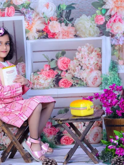 A girl in a stylish pink plaid outfit sits in a beautiful floral garden setup, reading a book. A sophisticated and sweet portrait for an older child.