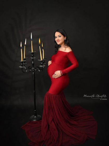 A dramatic and classic portrait. The mom-to-be stands elegantly in a red gown next to a candelabra prop, adding a touch of gothic romance to the scene.